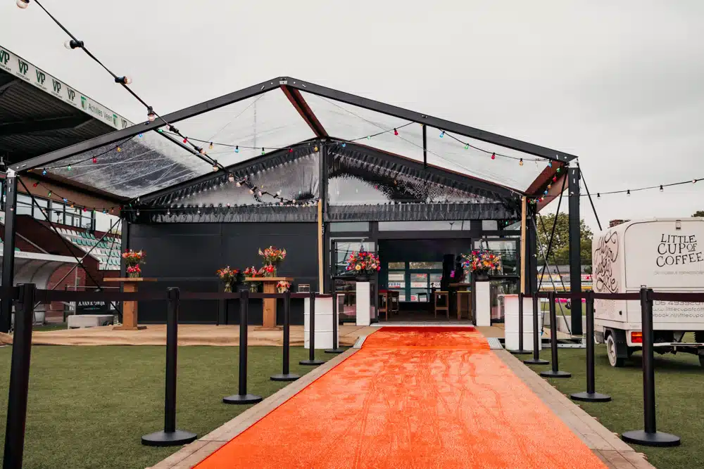 Luxe aluhal voor evenement met transparant dak, bloemen en opvallende oranje loper over sportveld