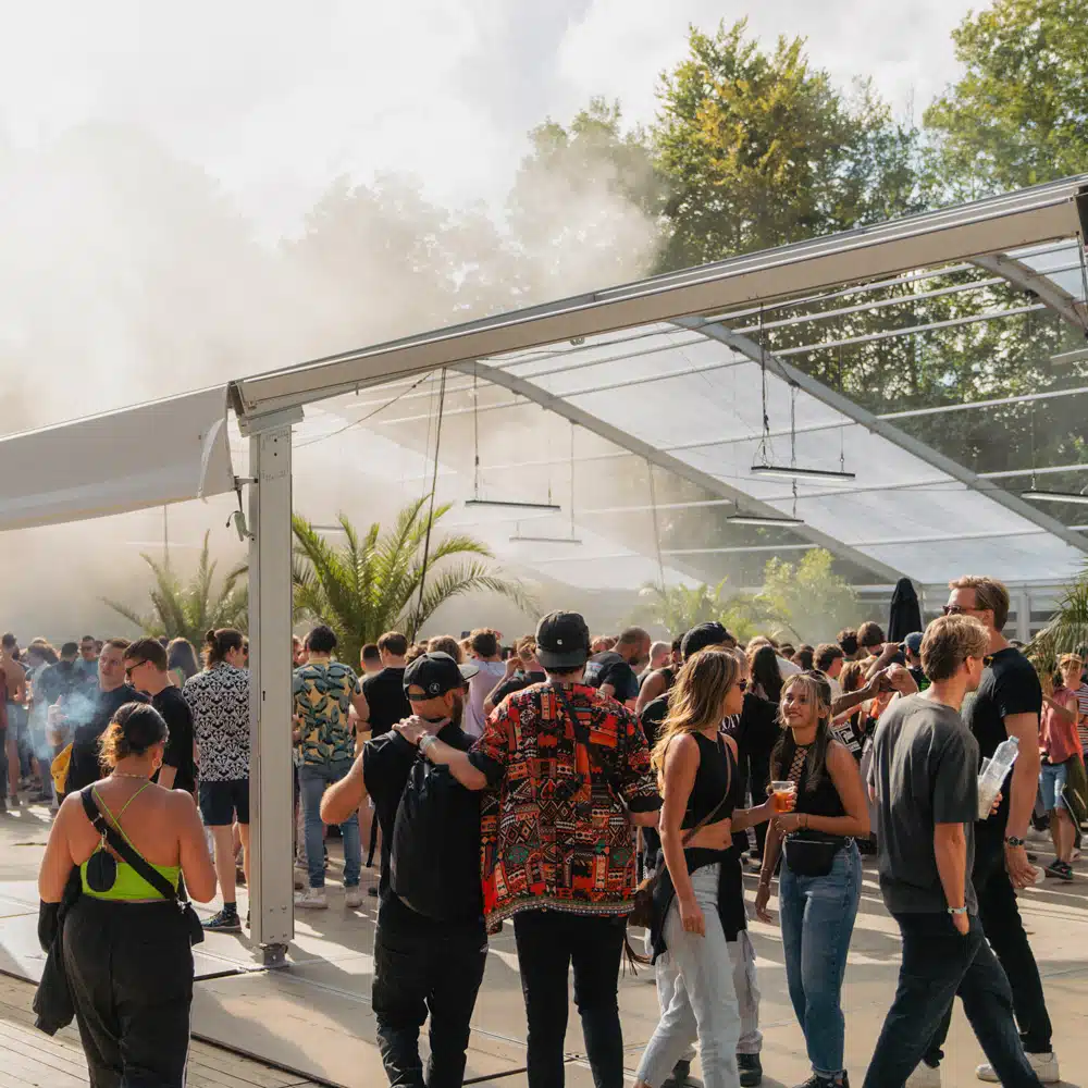 Transparante aluminium festivaltent tijdens een groot buiten muziekevenement met veel bezoekers.